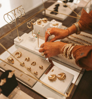 Woman's hands in One Dame Lane's retail store putting jewellery in the case. She is surrounded by displays of gold earrings, and is wearing bold bracelets and rings.
