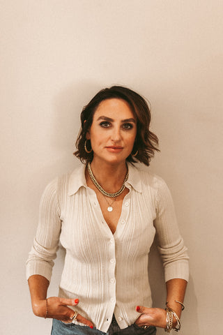 Image of One Dame Lane owner Lyndsey against a beige background. She is wearing a white top and blue jeans and has her hands in her pocket, and is wearing bold earrings, necklaces, bracelets, and rings.