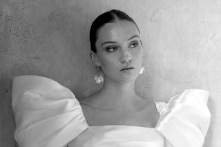 Black and white image of a woman against a textured background, wearing a white satin dress with puffed sleeves. She is wearing hoop earrings with pearl accents from ODL's Bridal collection.