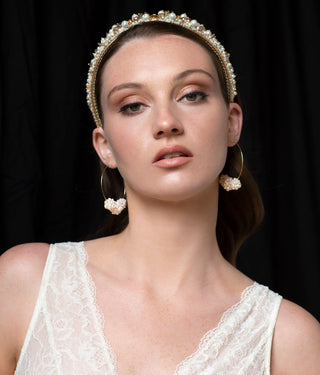 Woman posed on a black background wearing a white lace v-neck top. She is wearing gold and pearl hoop earrings and hair accessories from ODL's bridal collection.