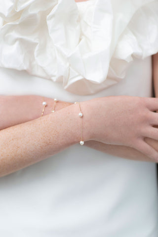 Woman in a white dress with her arms crossed, wearing delicate gold chain bracelets with pearls from One Dame Lane's bridal collection.