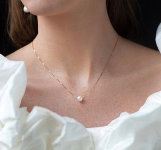 Woman posed against a black background wearing a frilly white top. She is wearing a delicate gold chain necklace with a pearl pendant from ODL's bridal collection.